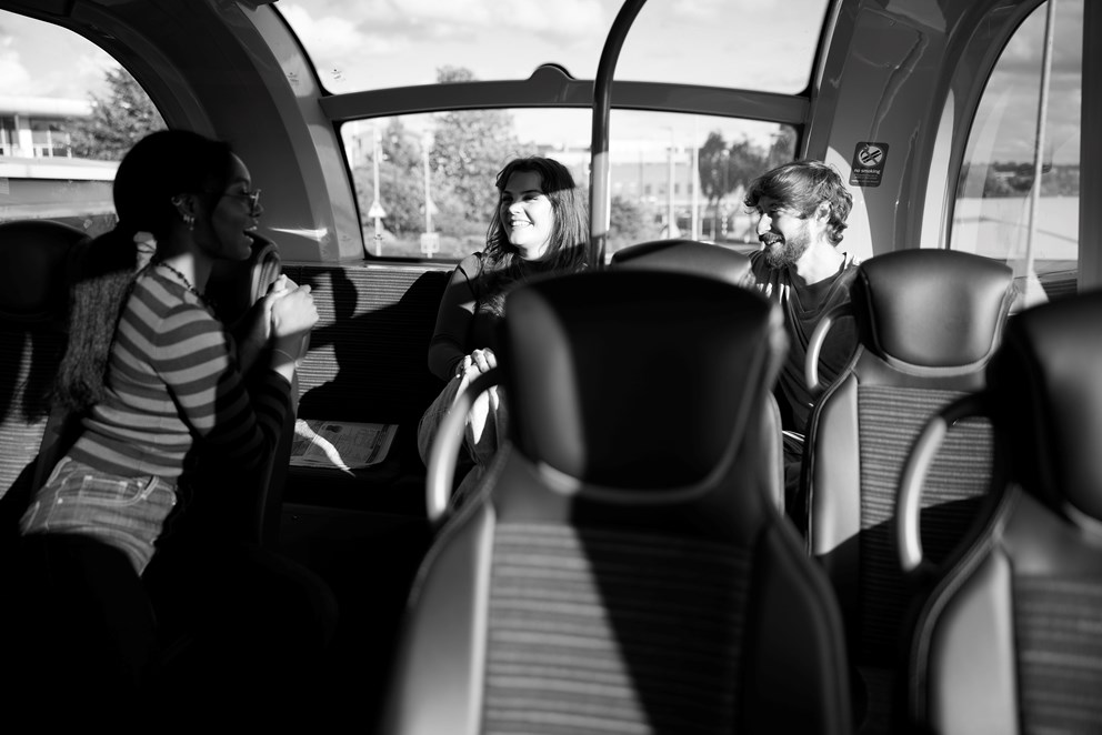 Three people sat at the back of a bus, talking and smiling.